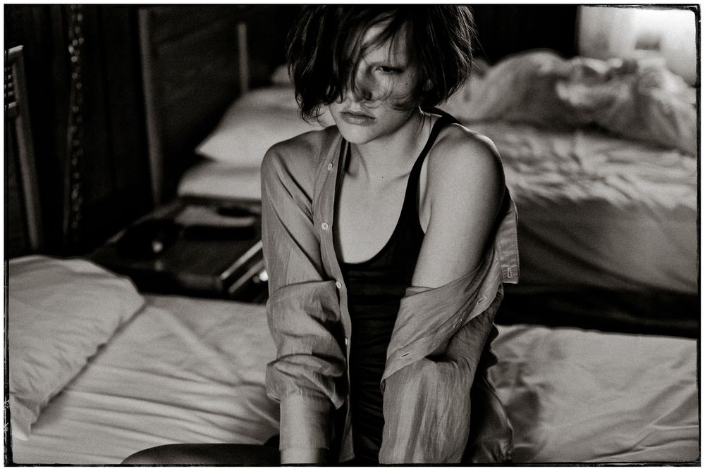 Black and white photograph of a woman in a tank top sitting on a bed in a dimly lit bedroom.