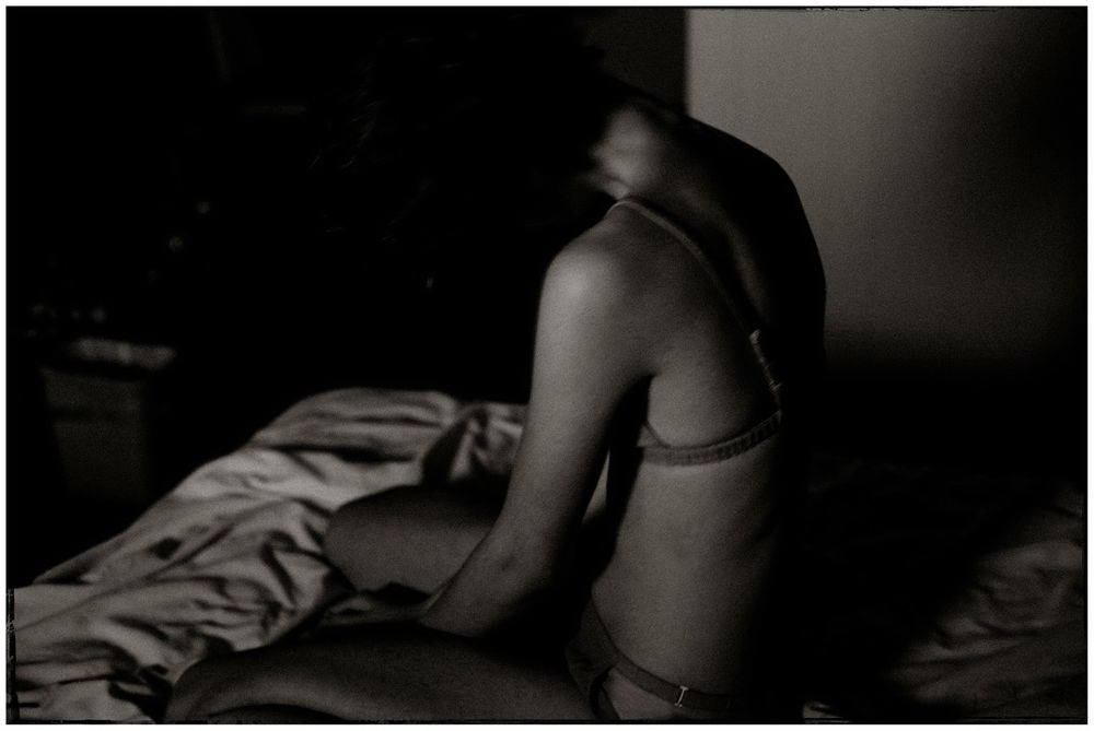 Black and white photo of a woman sitting on a bed in lingerie, her face blurred in shadow.