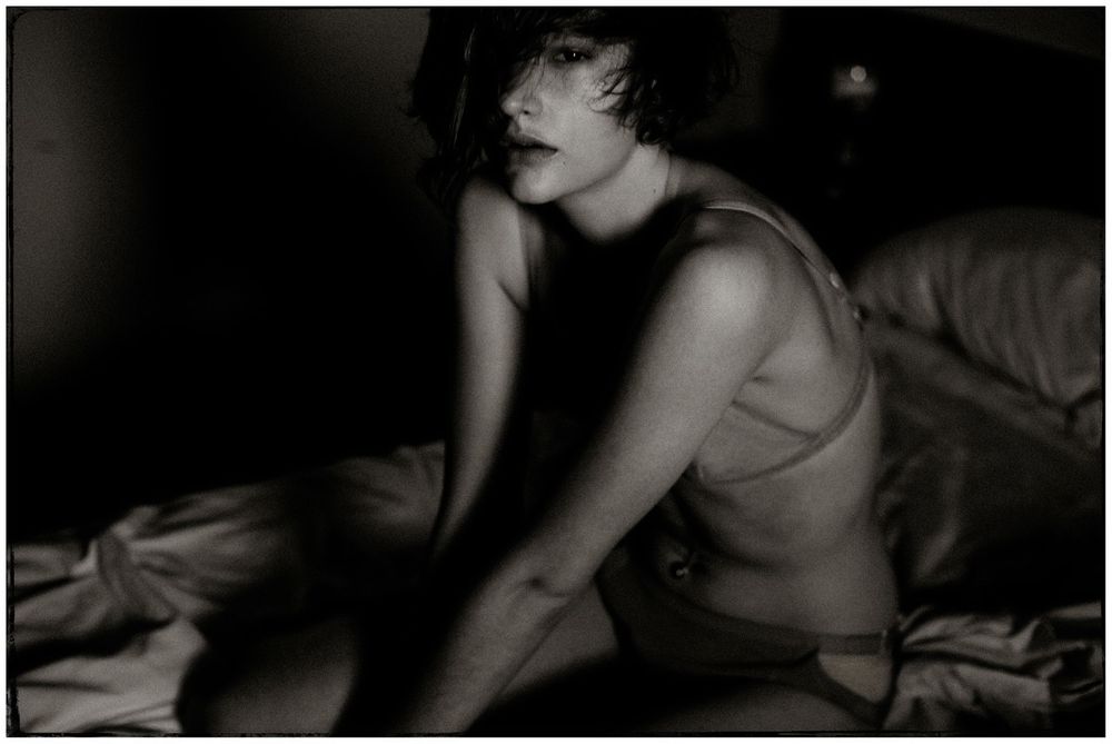 Black and white photograph of a woman in a tank top sitting on a bed in dim lighting.