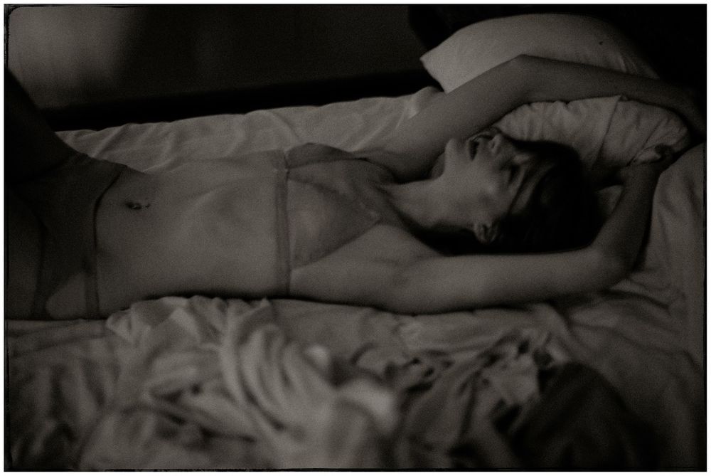 Black and white artistic photo of a woman in sheer lingerie lying on a bed with rumpled sheets.