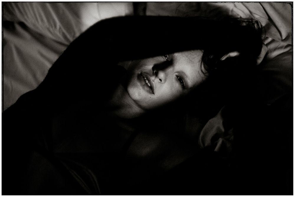 Black and white portrait of a smiling woman lying on a bed with her arms raised above her head.