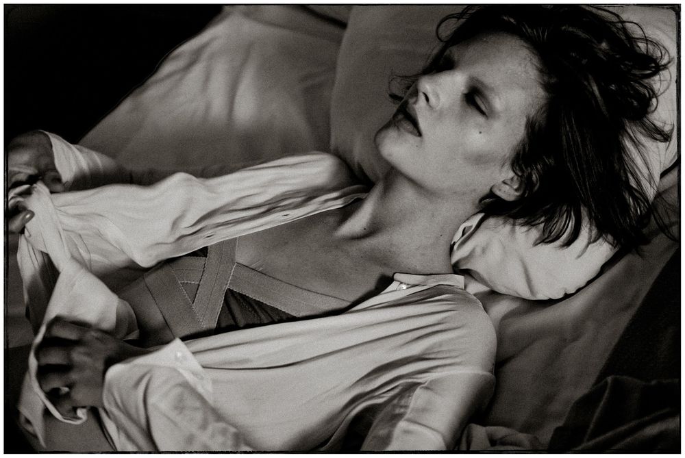 Black and white photo of a young person reclining dramatically in an open white shirt with tousled dark hair.