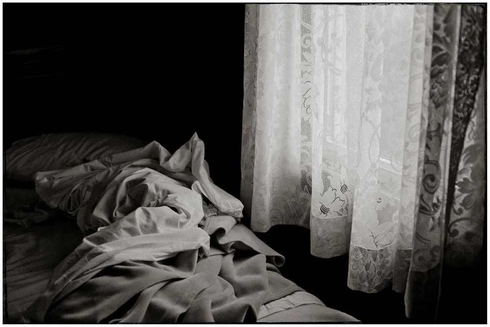 Black and white photograph of an unmade bed with rumpled sheets near a window with sheer curtains in dim lighting.