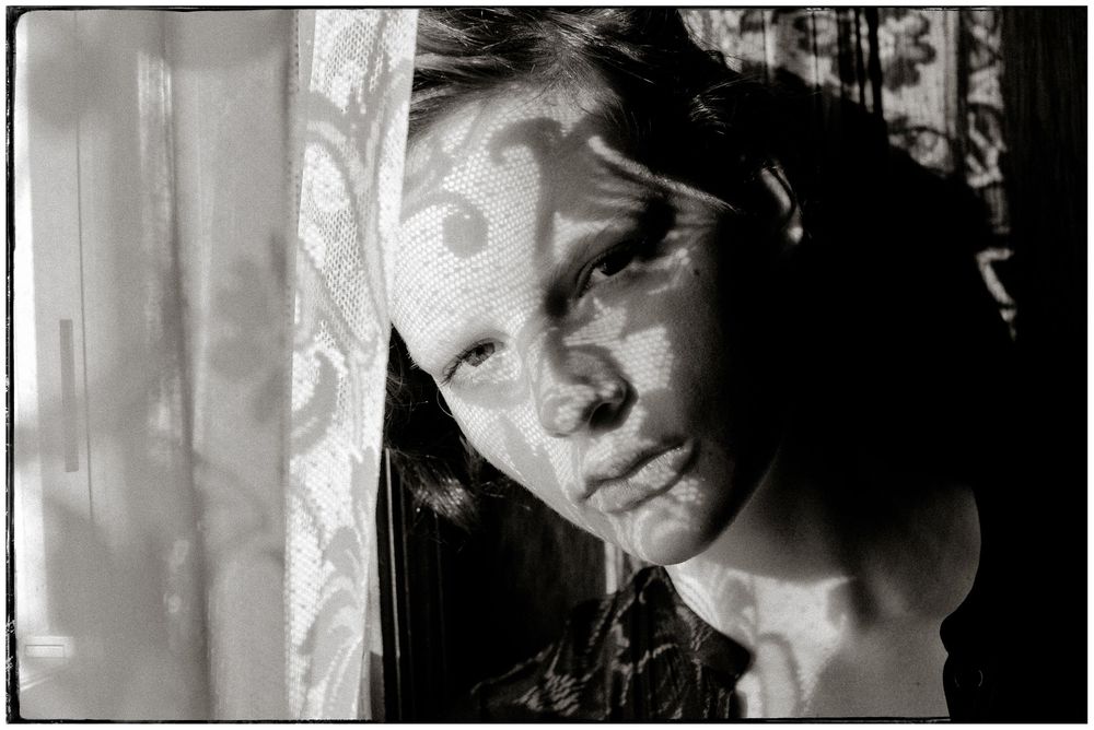 Black and white portrait of a woman with dramatic lighting through window curtains creating shadows across her face.