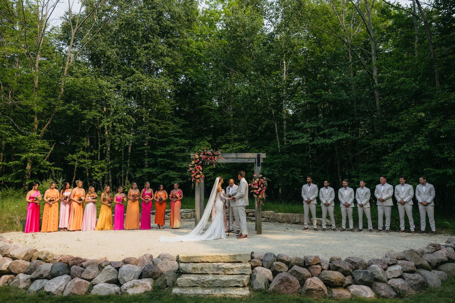 Wedding ceremony in a forest setting with wedding party lined up on stone platform surrounded by lush greenery.