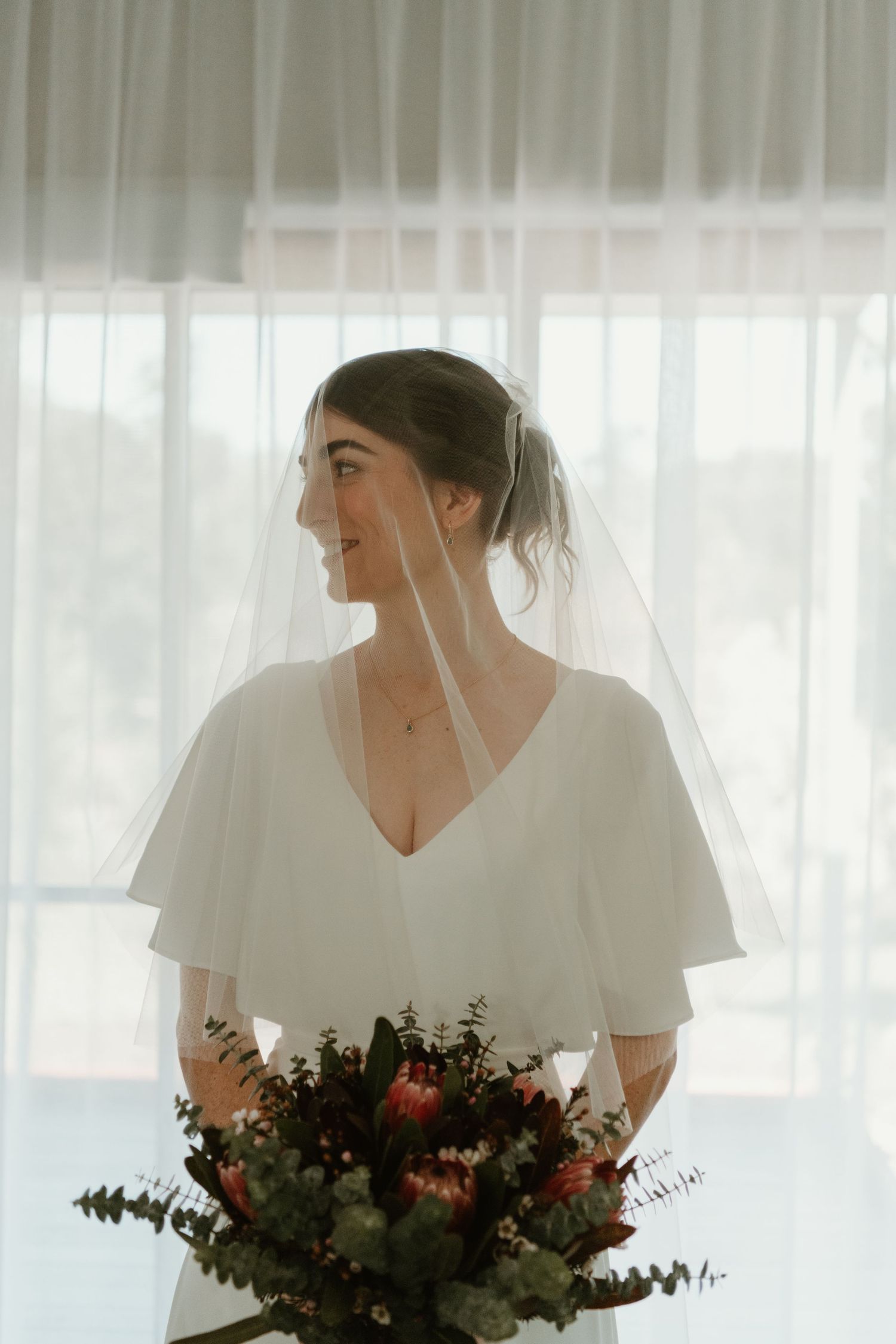 Bride in white flutter-sleeve gown and veil holds a bouquet of proteas, smiling softly by a bright window.
