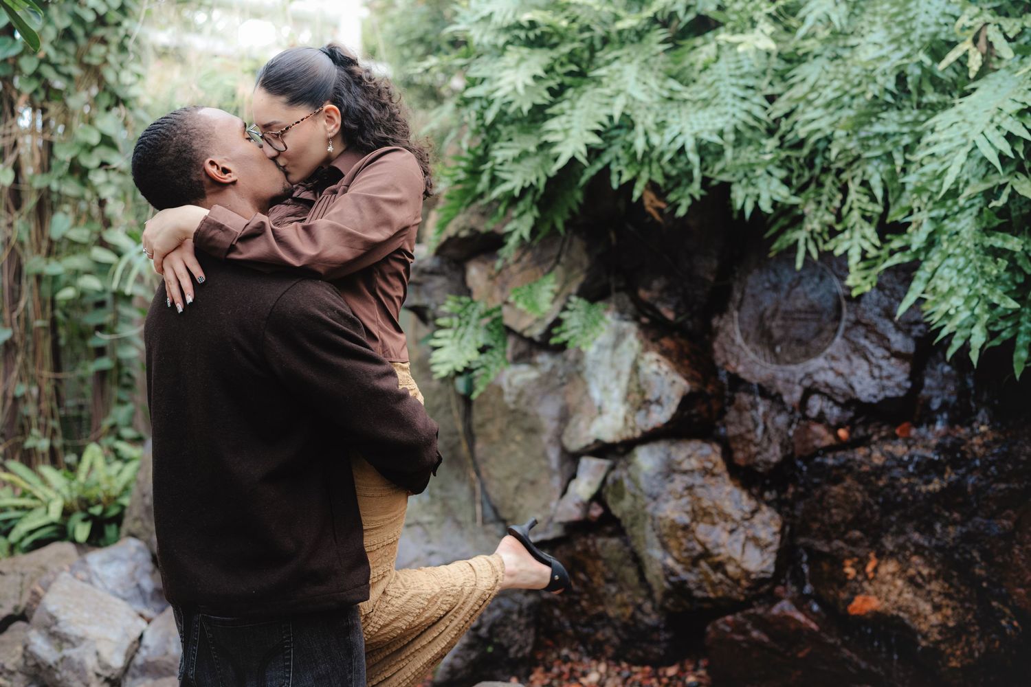 Couple sharing a romantic kiss near a rocky waterfall surrounded by green ferns.