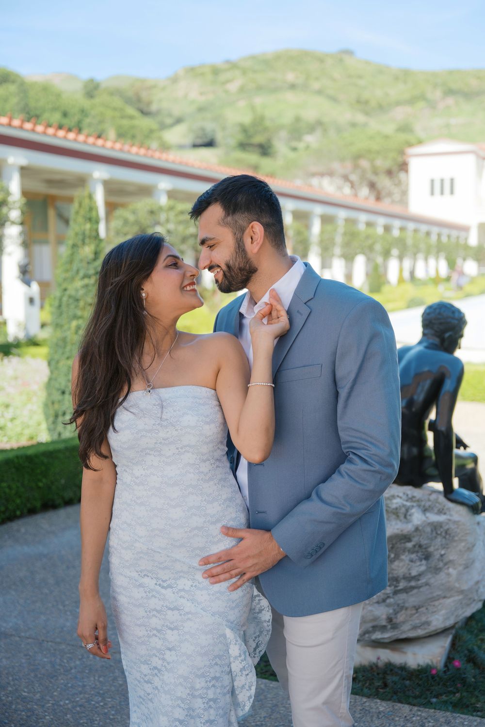 A couple shares a romantic moment in formal attire at an outdoor venue with hills in the background.