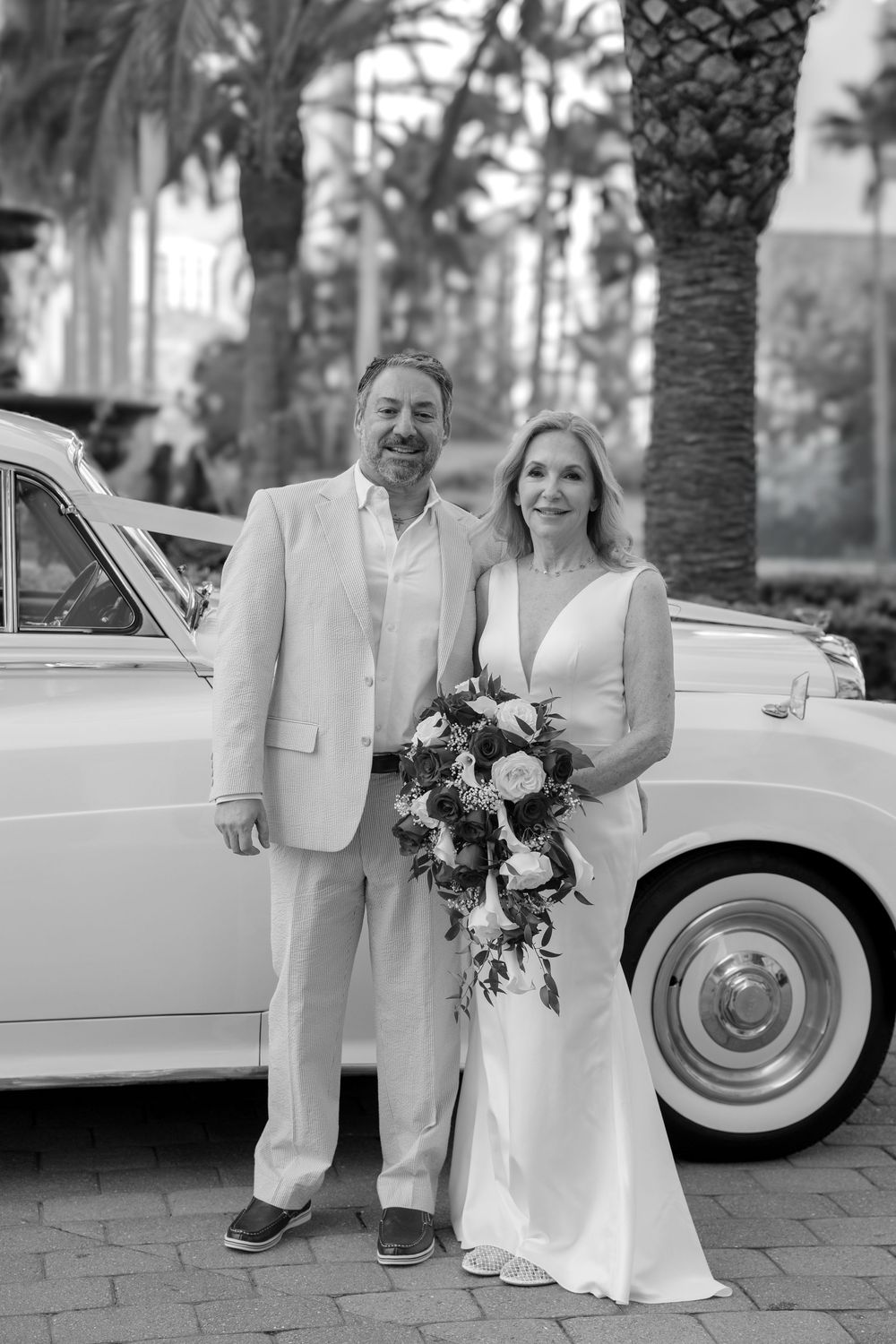 A bride and groom pose in black and white beside a vintage white car with palms.
