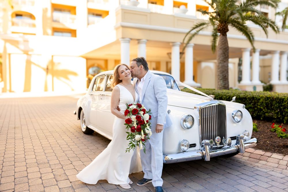 A bride and groom kiss beside a classic white Rolls-Royce with red rose bouquet.