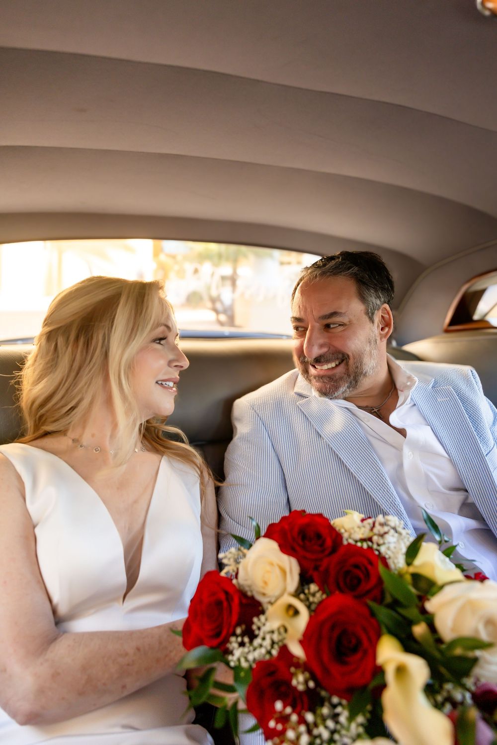 A bride and groom smile at each other in a vintage car with a red rose bouquet.