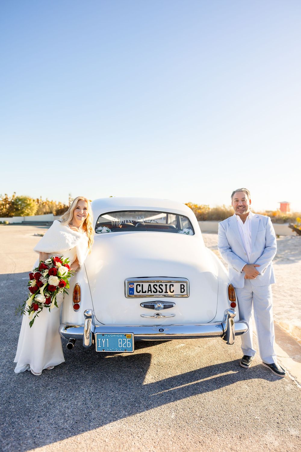 Bride and groom posing behind a classic white vintage car with CLASSIC B license plate on a sunny day.