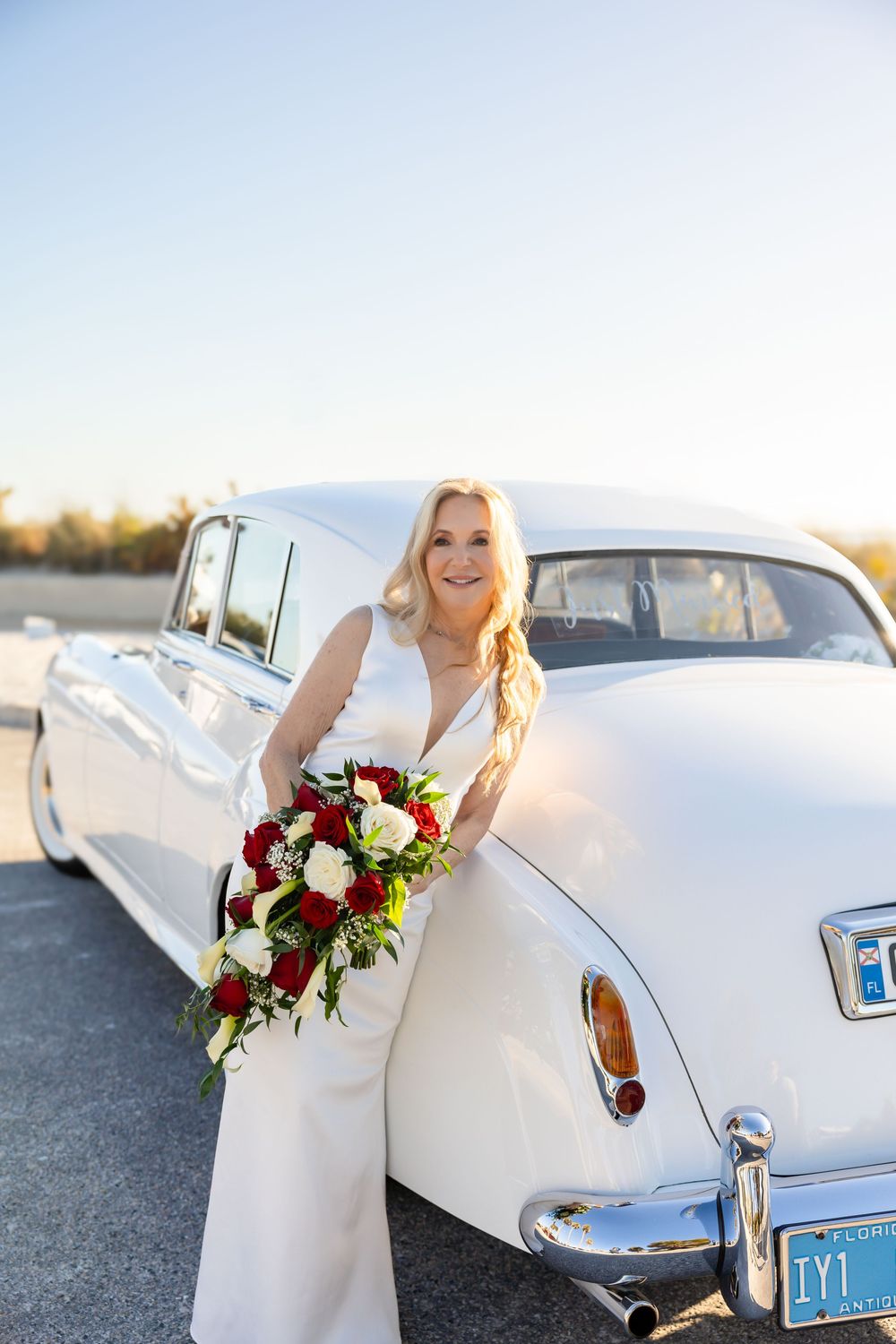 Elegant blonde bride in white gown holding red and white rose bouquet leaning against a classic white car.