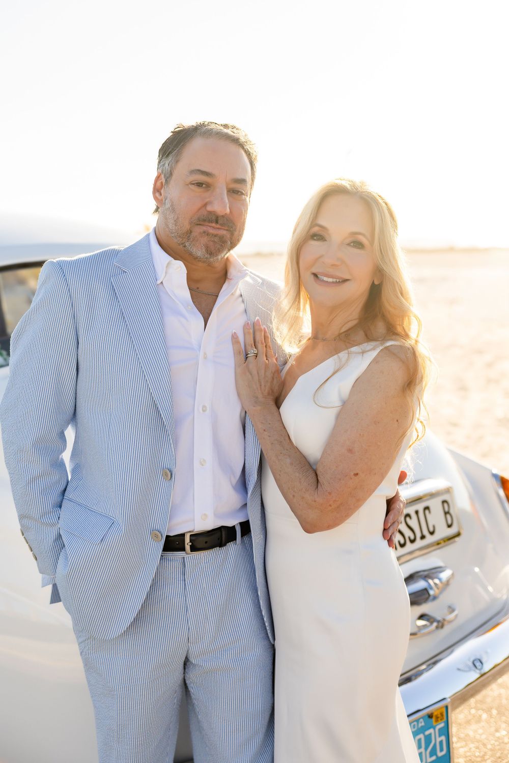 Smiling wedding couple posing together in front of a vintage white car at golden hour sunset.
