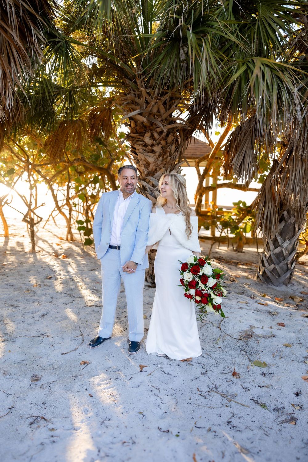 Bride and groom holding hands under a tropical palm tree on sandy beach with red rose bouquet.