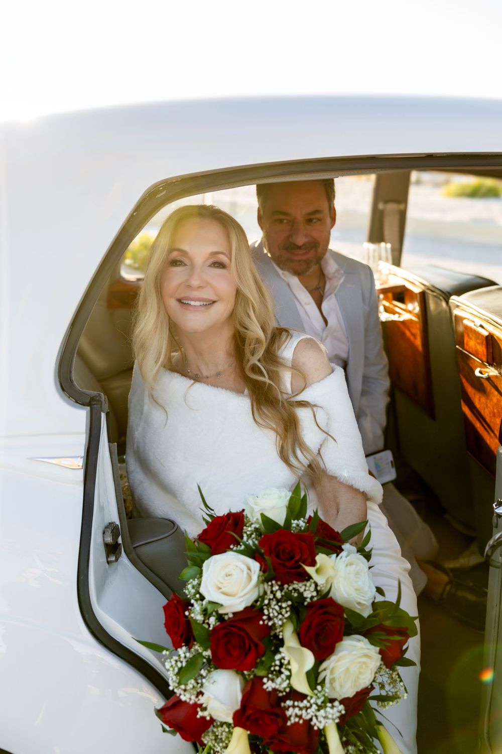 Radiant blonde bride holding red and white rose bouquet stepping out of a classic white vintage car.