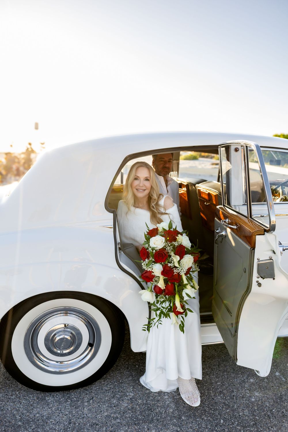 Smiling bride in white dress holding cascading red and white rose bouquet seated in vintage white car.