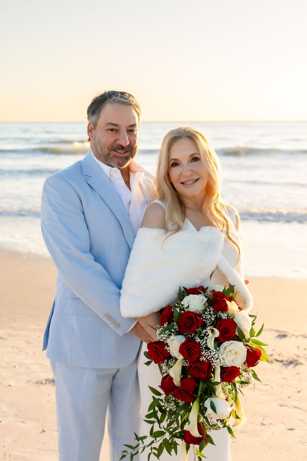 Bride and groom embracing on a sandy beach at sunset with red and white rose bouquet.