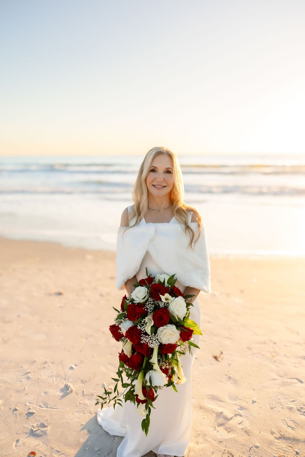 Bride in white dress and wrap holds red and white bouquet on a sunny beach at sunset.