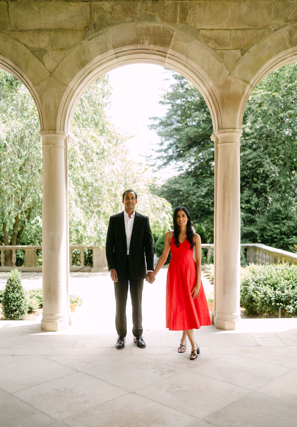 A romantic couple stands beneath elegant stone arches in a garden setting, creating a sequence of artistic poses.