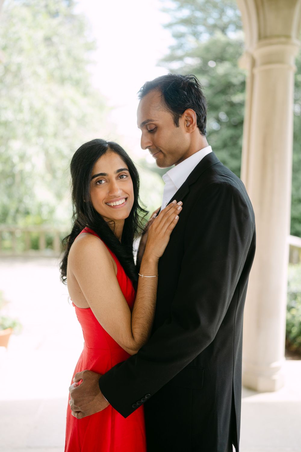 A couple in formal attire embrace on an elegant porch, with the woman wearing a vibrant red dress and the man in a black suit.