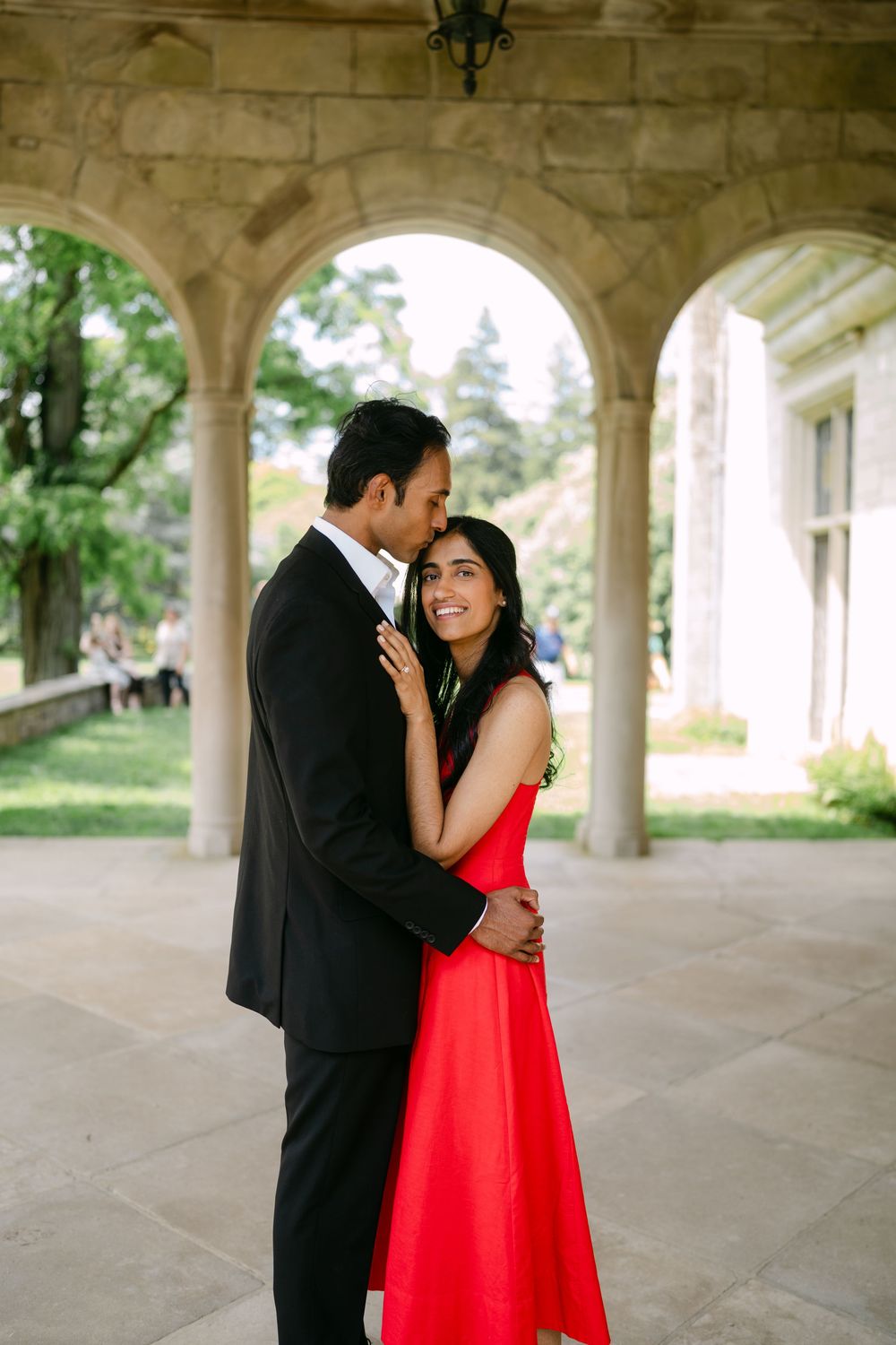 A couple shares an intimate moment under stone archways, with the person in red wearing a vibrant dress.