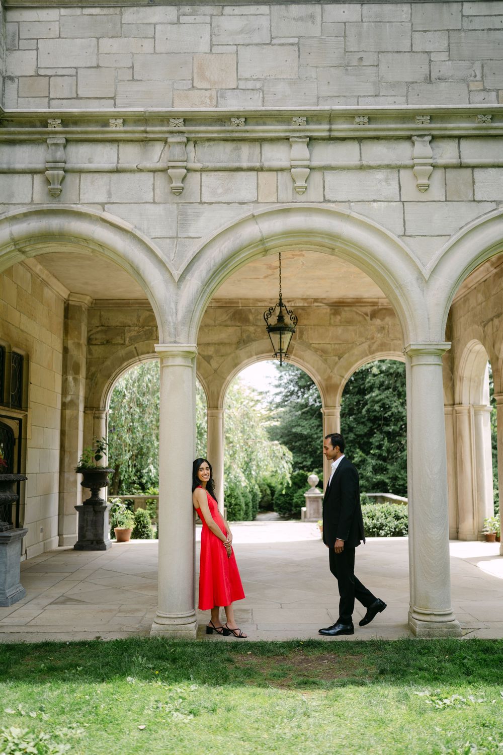 Couple in formal attire poses under elegant stone archways surrounded by greenery during a romantic photoshoot.