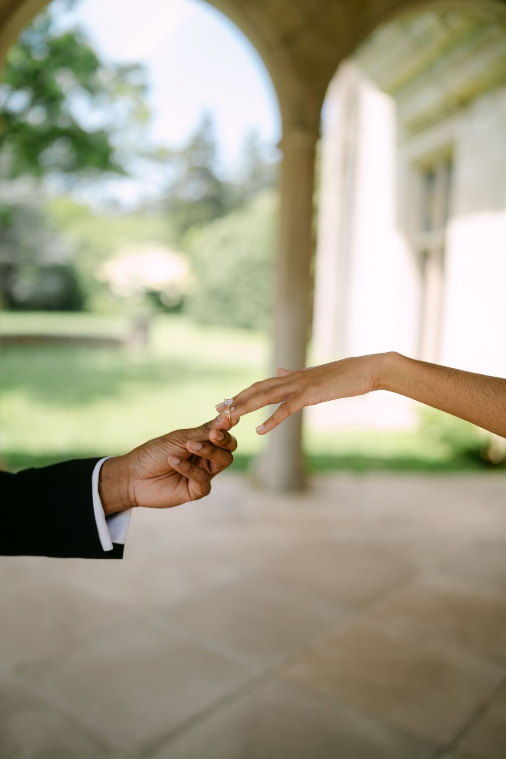 Two hands reach towards each other under an arched colonnade with sunlit garden in background.