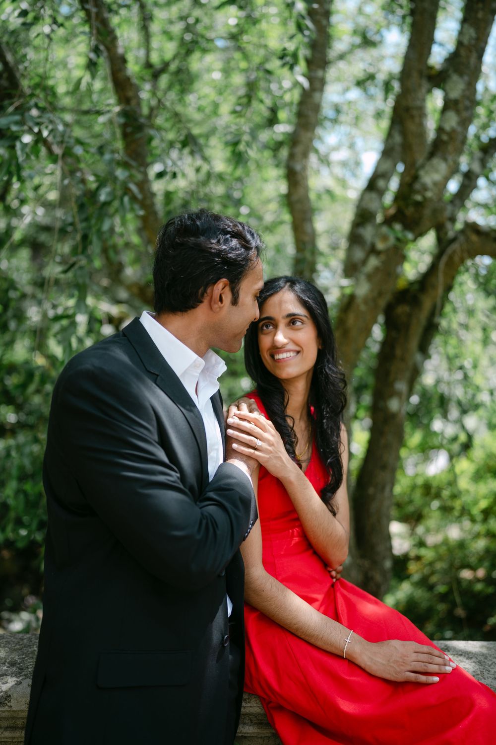 A romantic couple embraces in a wooded setting, with the woman wearing a vibrant red dress against lush greenery.