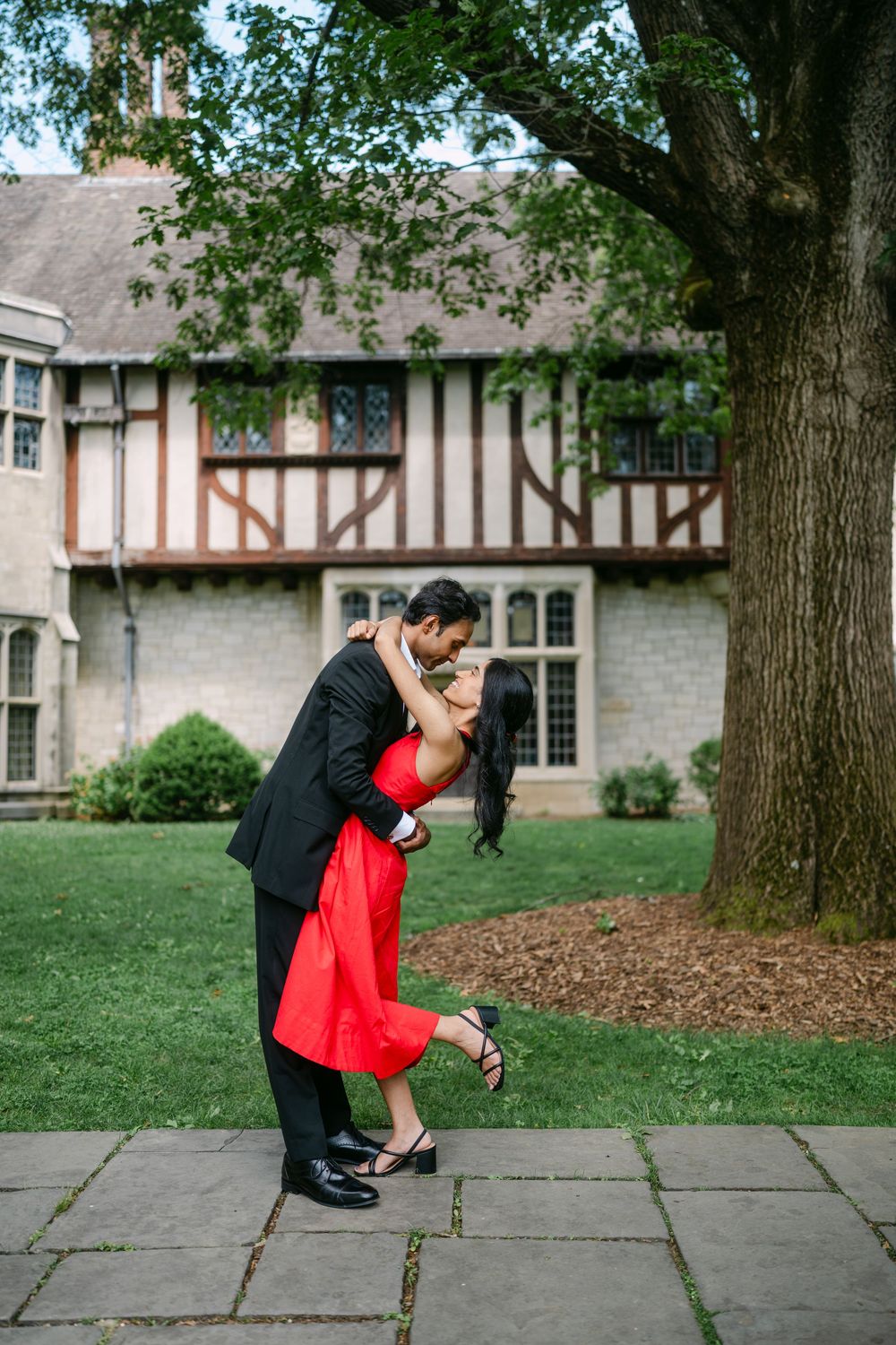 A couple in formal attire shares a romantic moment on stone pavement near a Tudor-style building.