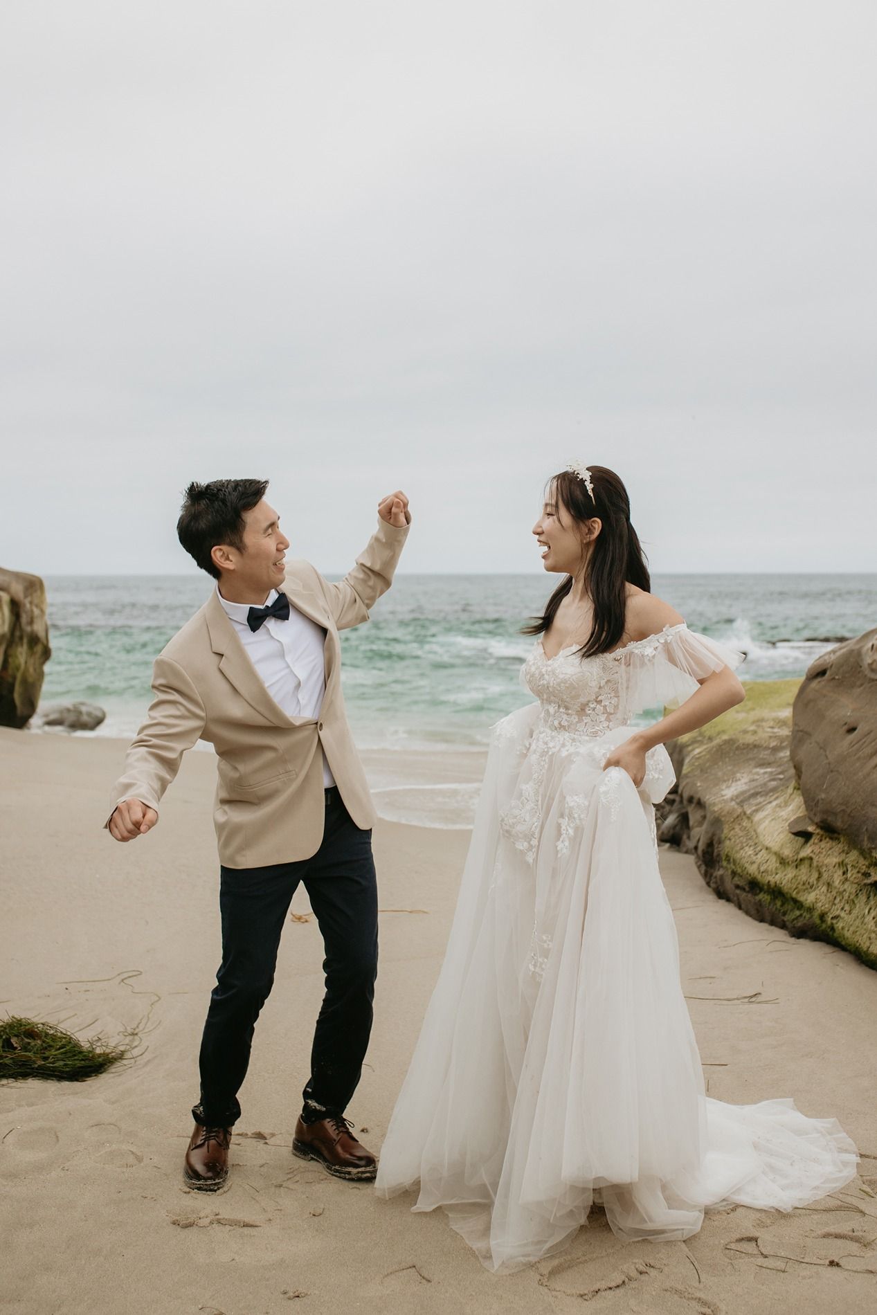 A couple in wedding attire dances playfully together on a beach with ocean waves in the background at Windansea Beach in La Jolla.