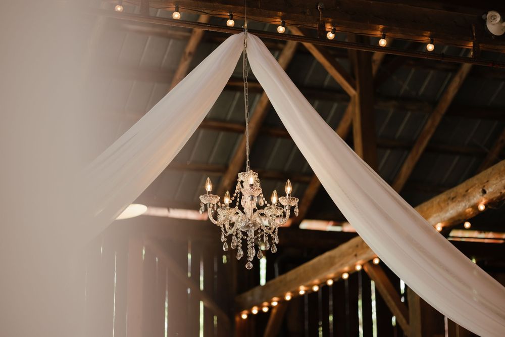 An elegant crystal chandelier hangs beneath white fabric draping and string lights in a rustic wooden barn ceiling.