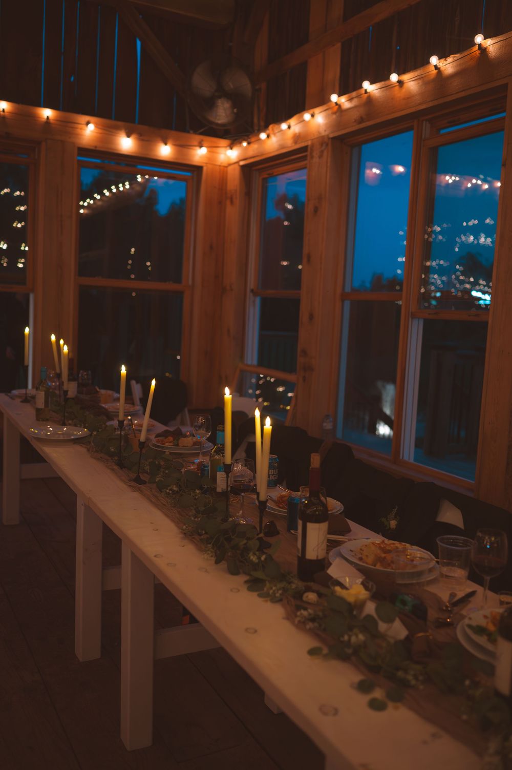 String lights illuminate a cozy indoor dining area with large windows overlooking a nighttime city view.