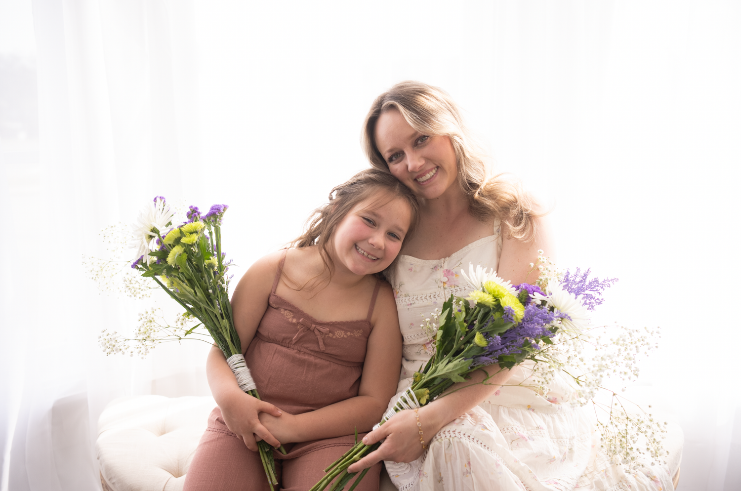 Two women in neutral tones pose together with flowers in a bright, airy studio setting.