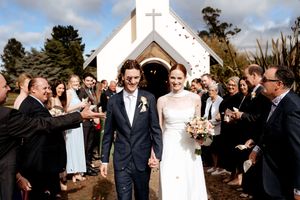 Wedding couple exits white chapel while guests throw confetti and celebrate along the pathway against cloudy sky backdrop.