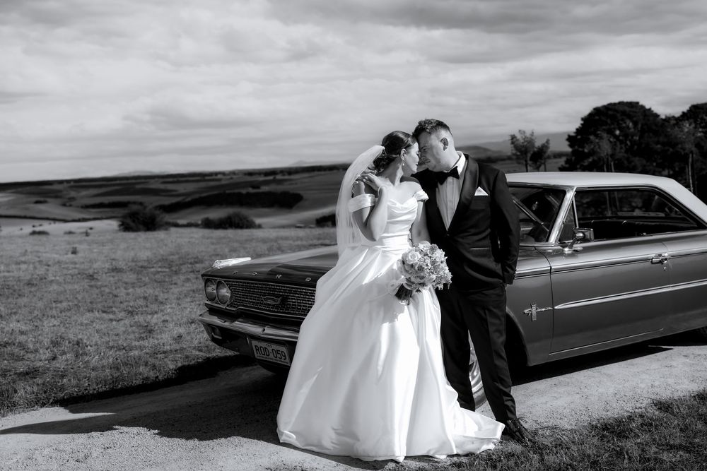 A couple in wedding attire stands beside a classic car on a hilltop overlooking a scenic landscape in black and white.