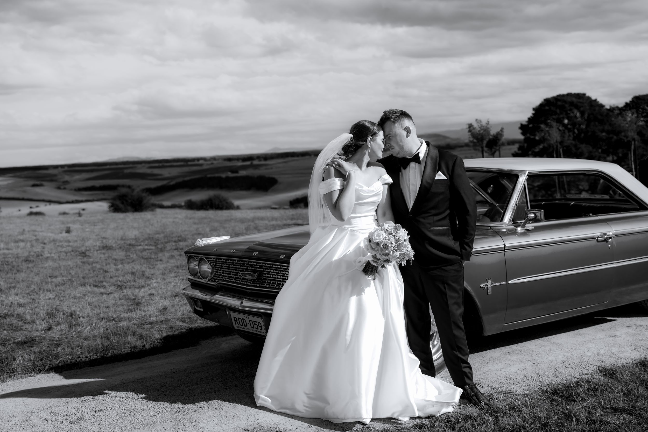 A couple in wedding attire stands beside a classic car on a hilltop overlooking a scenic landscape in black and white.