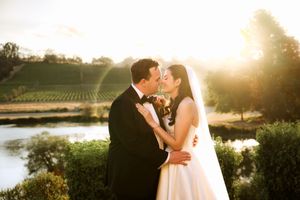 A couple shares an intimate moment at sunset by a vineyard and lake in formal wedding attire.