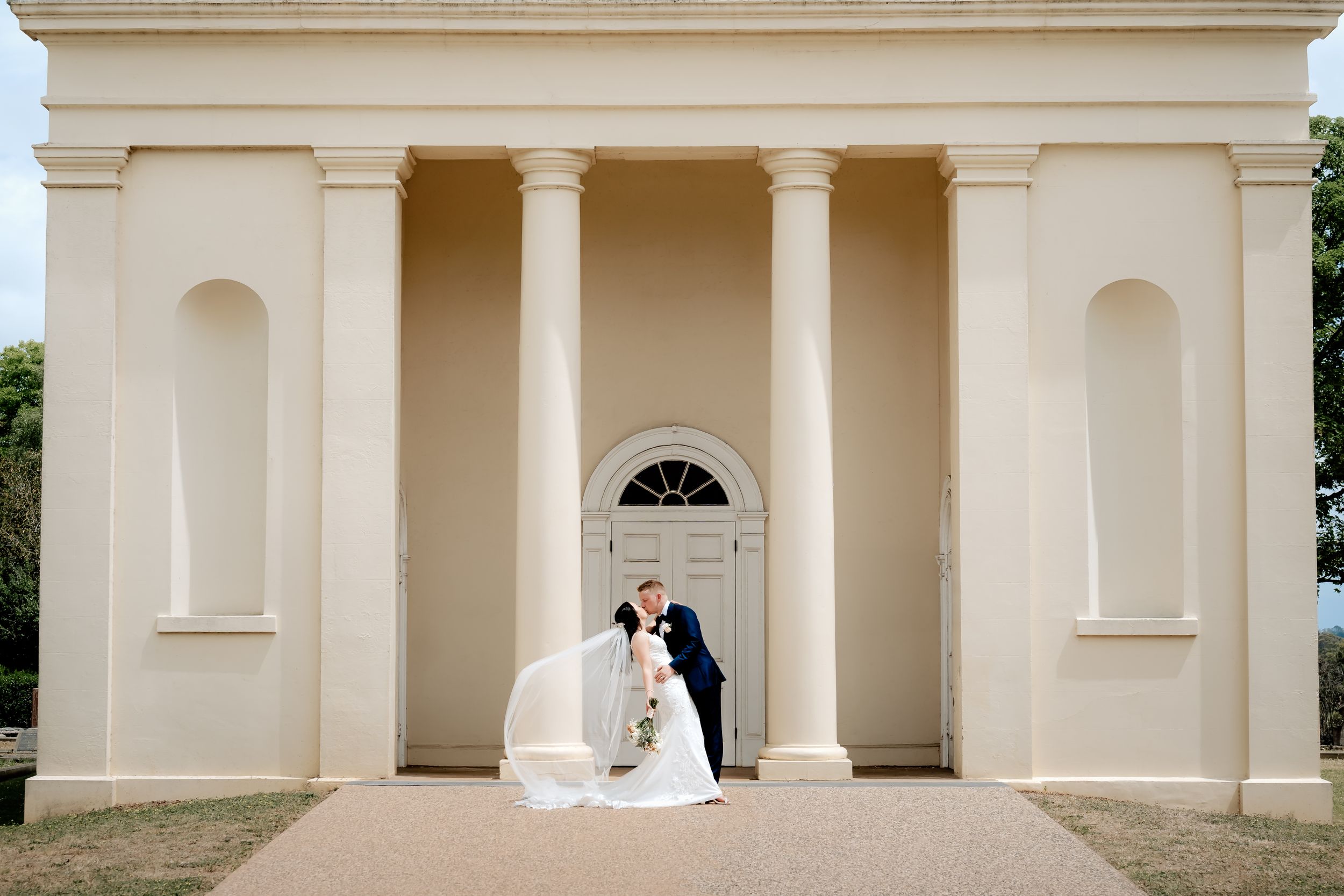 Elegant wedding couple poses in front of a classical columned building entrance with cream colored walls.