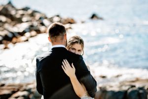 Wedding couple embraces on rocky coastline with sparkling ocean in background.