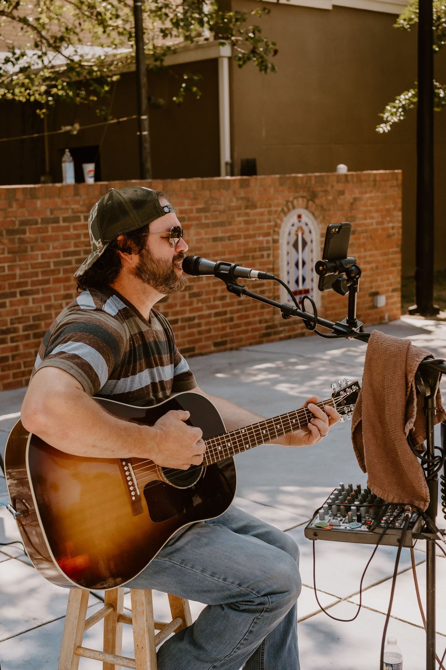 A musician performs outdoors with an acoustic guitar and microphone setup on a sunny patio near a brick wall.