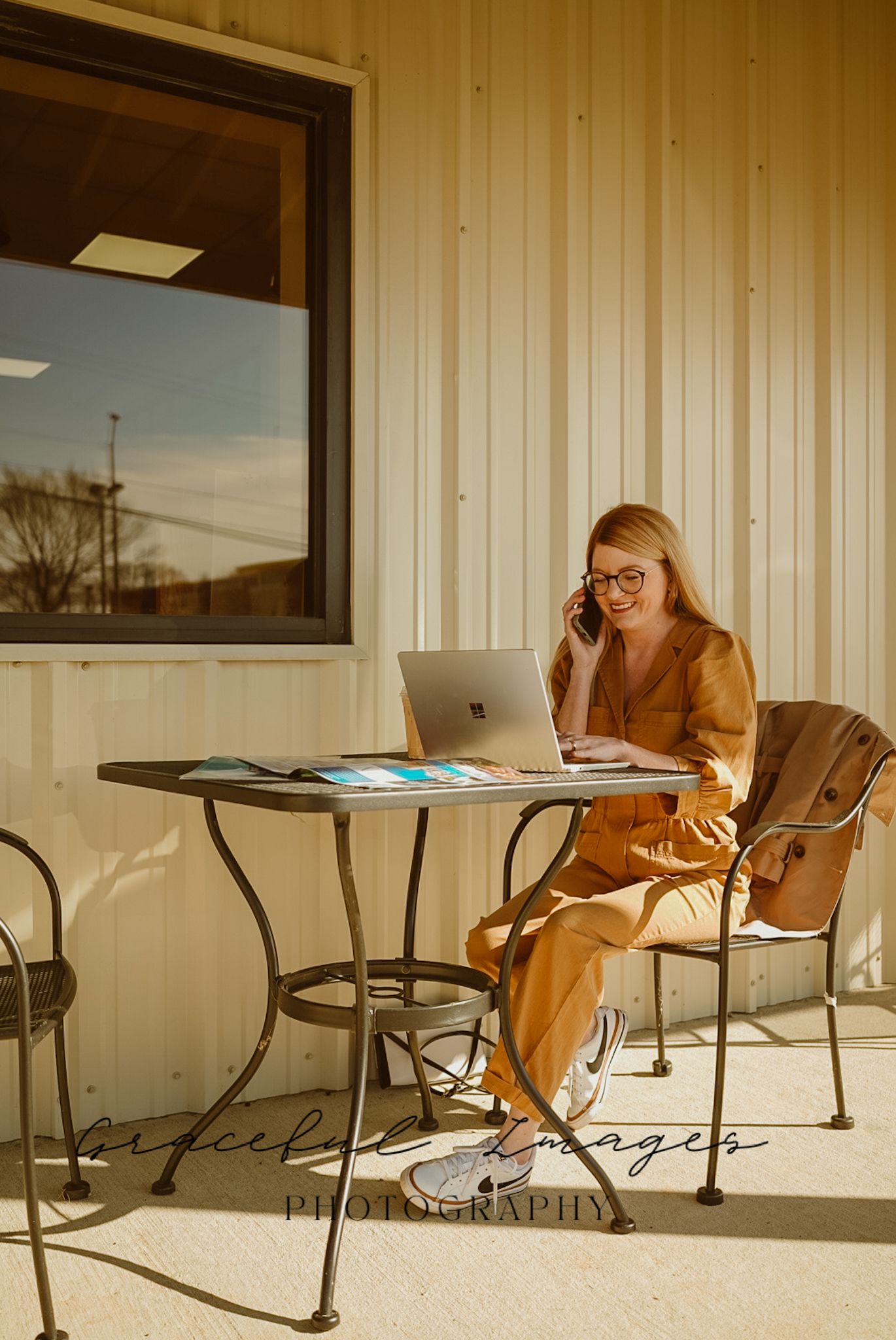 A person in a mustard yellow dress works on a laptop at an outdoor metal table in warm sunlight.