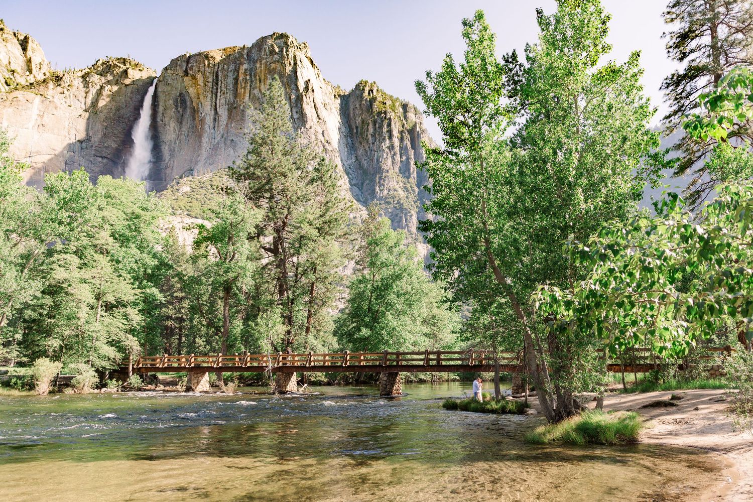 Scenic view of Yosemite Valley with waterfall, cliffs and wooden footbridge in the distance.