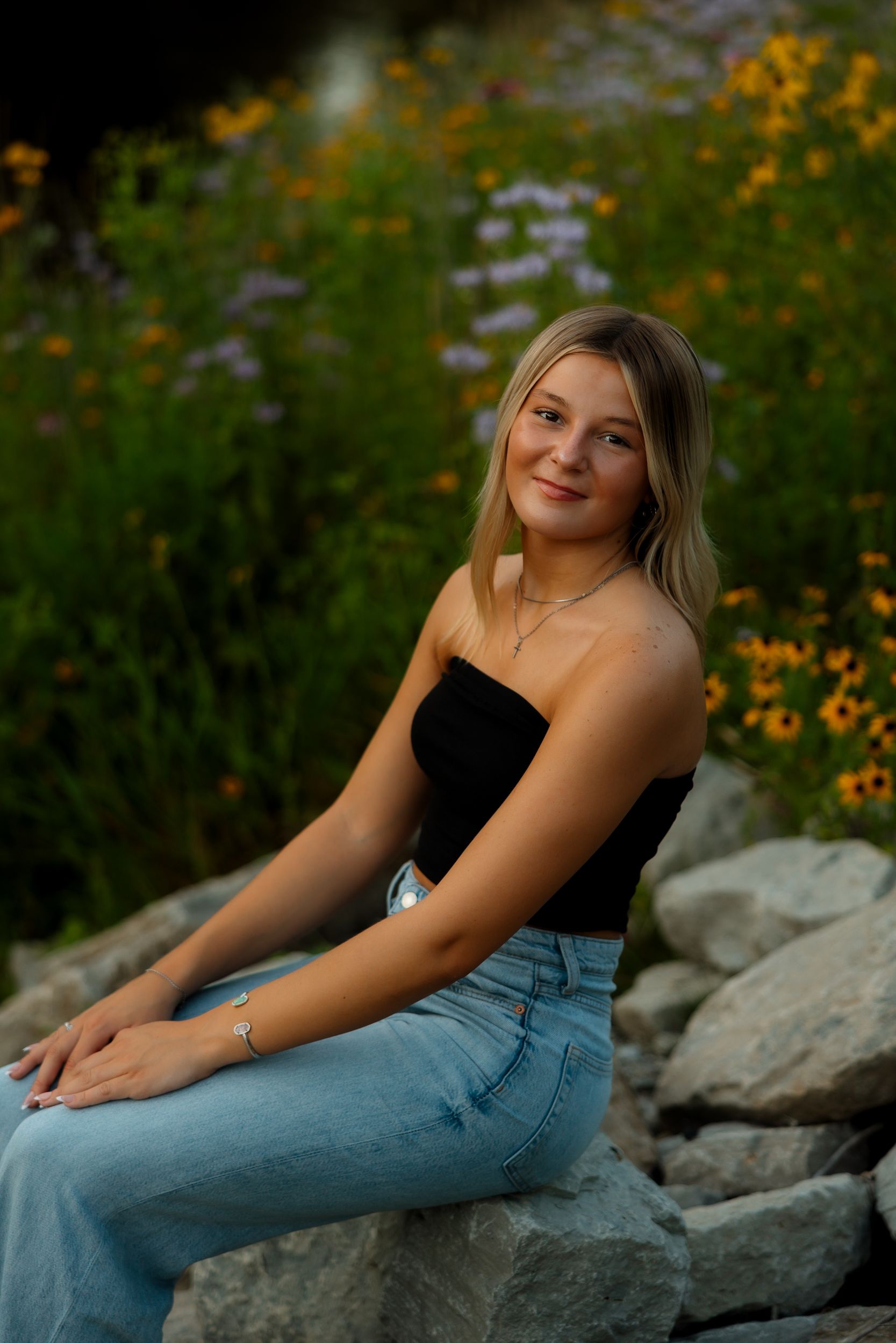 Woman in casual attire poses on rocky terrain surrounded by vibrant yellow wildflowers in a natural outdoor setting.