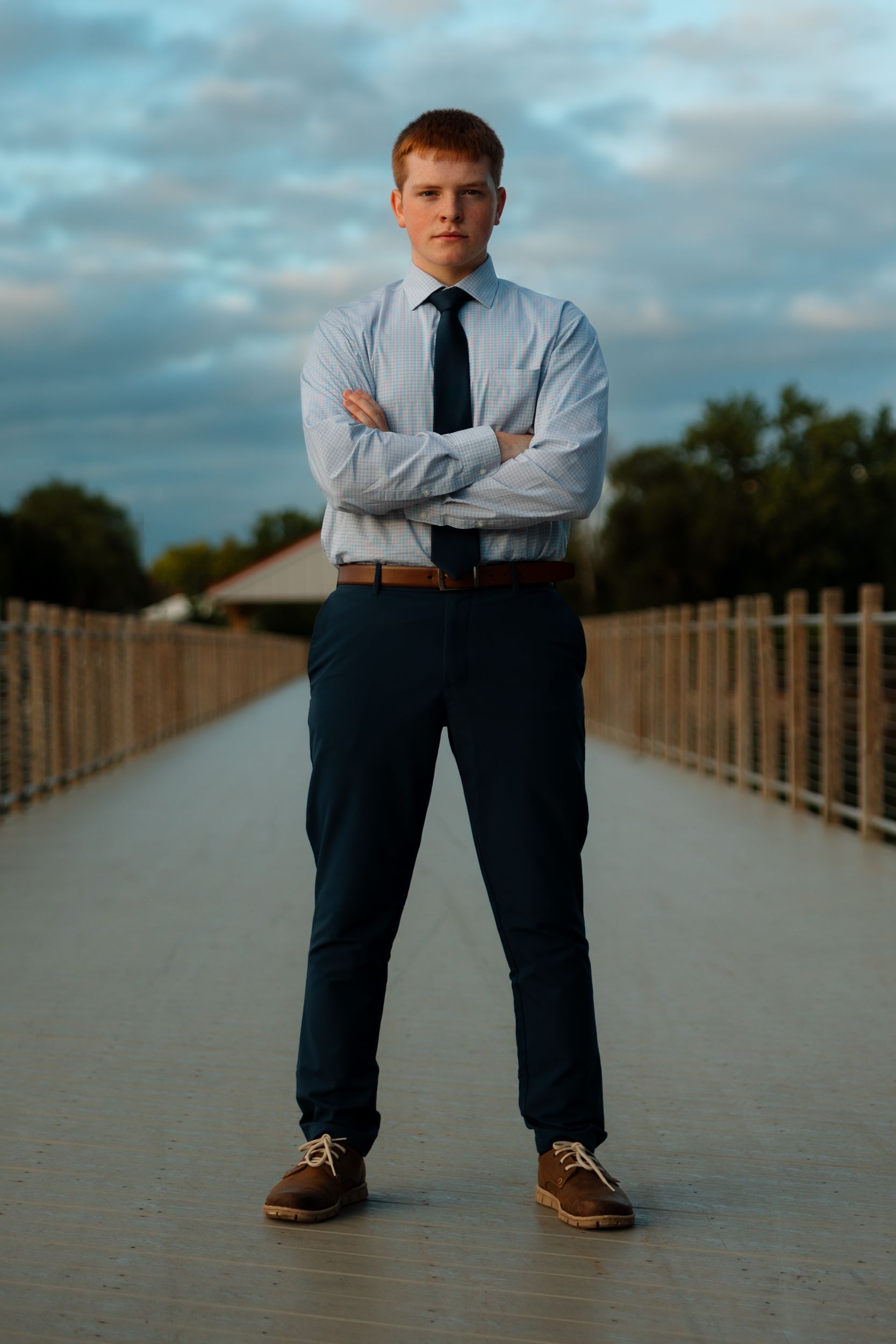 A person in business attire stands on a wooden boardwalk with arms crossed during an outdoor photoshoot.