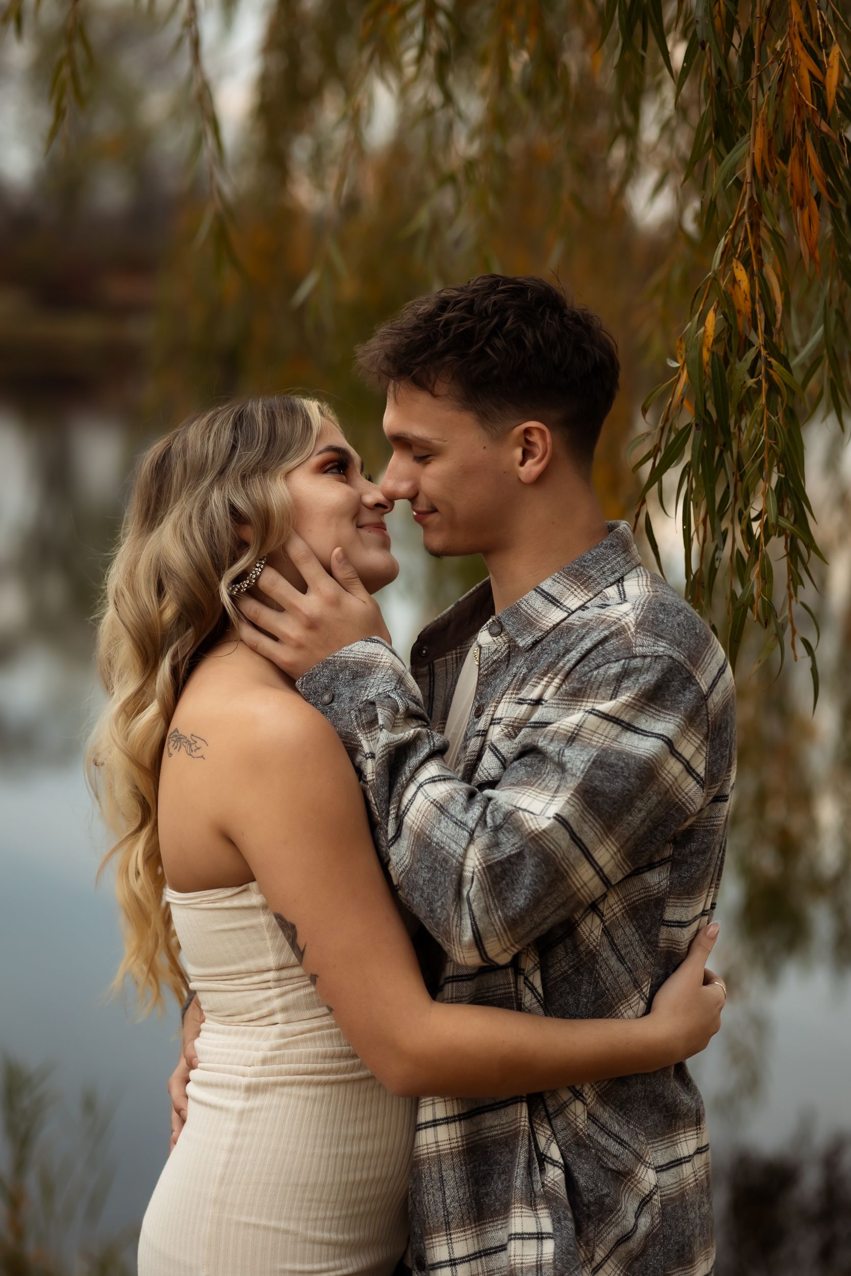 A romantic couple embracing under willow branches near water during golden hour.