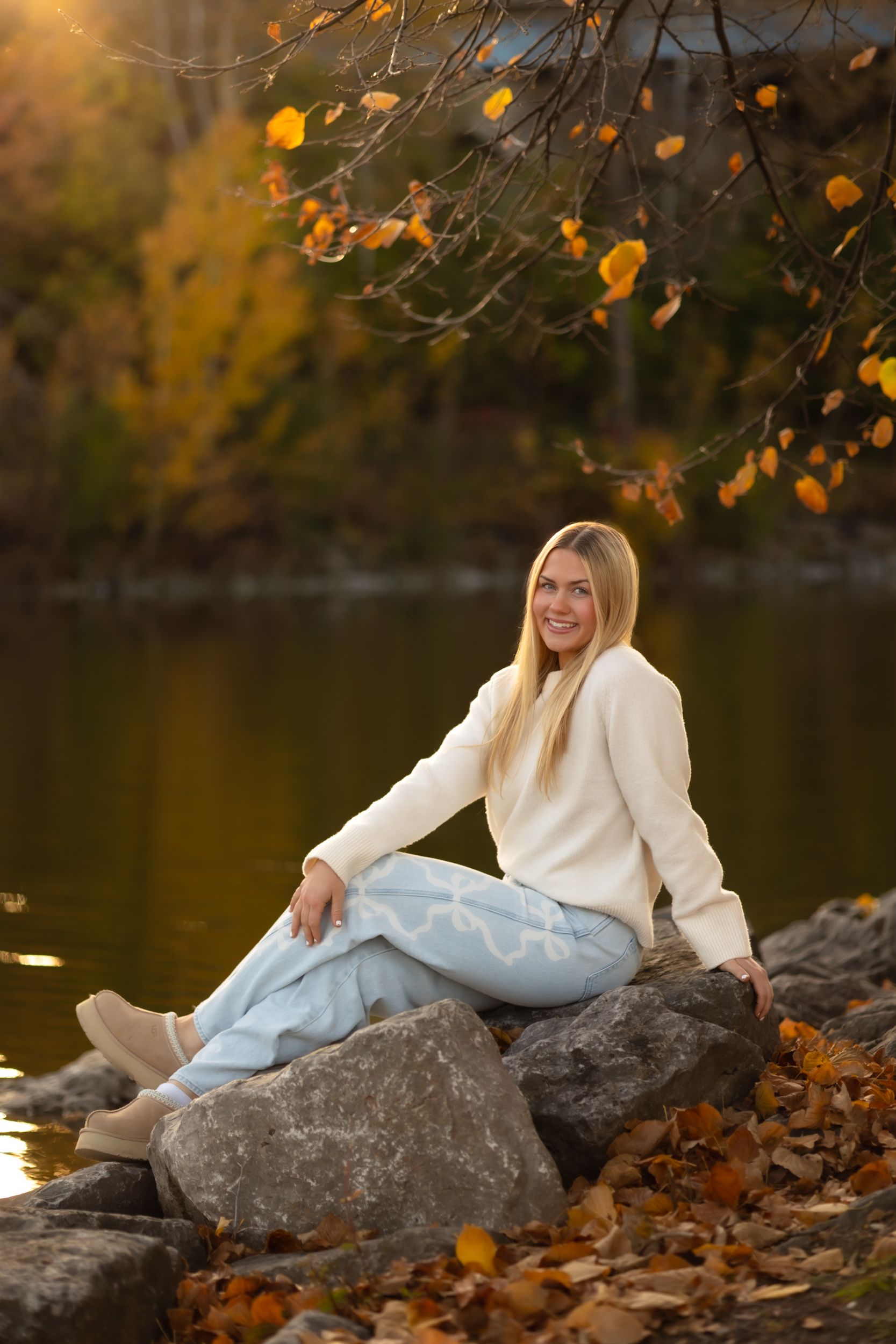 A person in a white sweater reclines on large rocks by a lake surrounded by autumn foliage and yellow leaves.