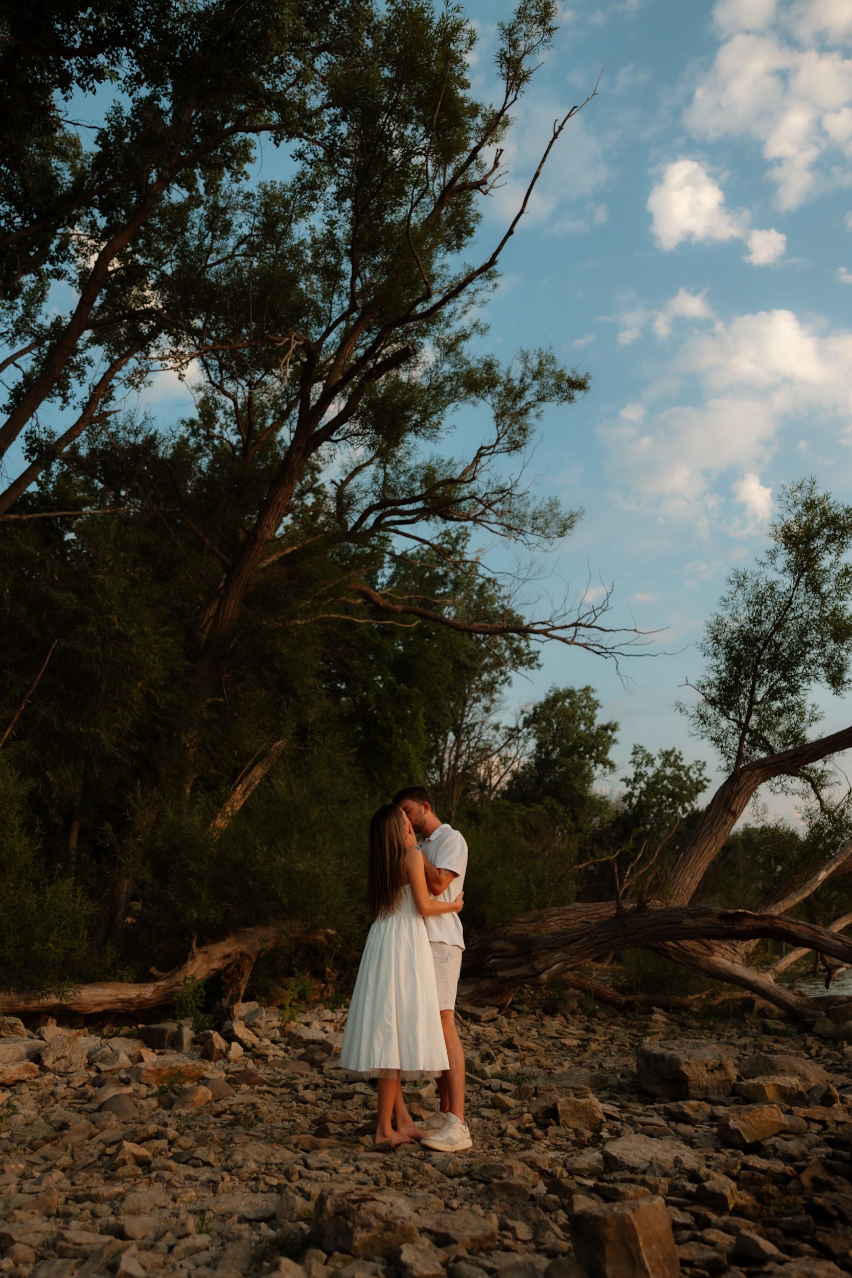 A romantic sunset moment captured on a rocky beach with tall trees and fluffy clouds in the background.
