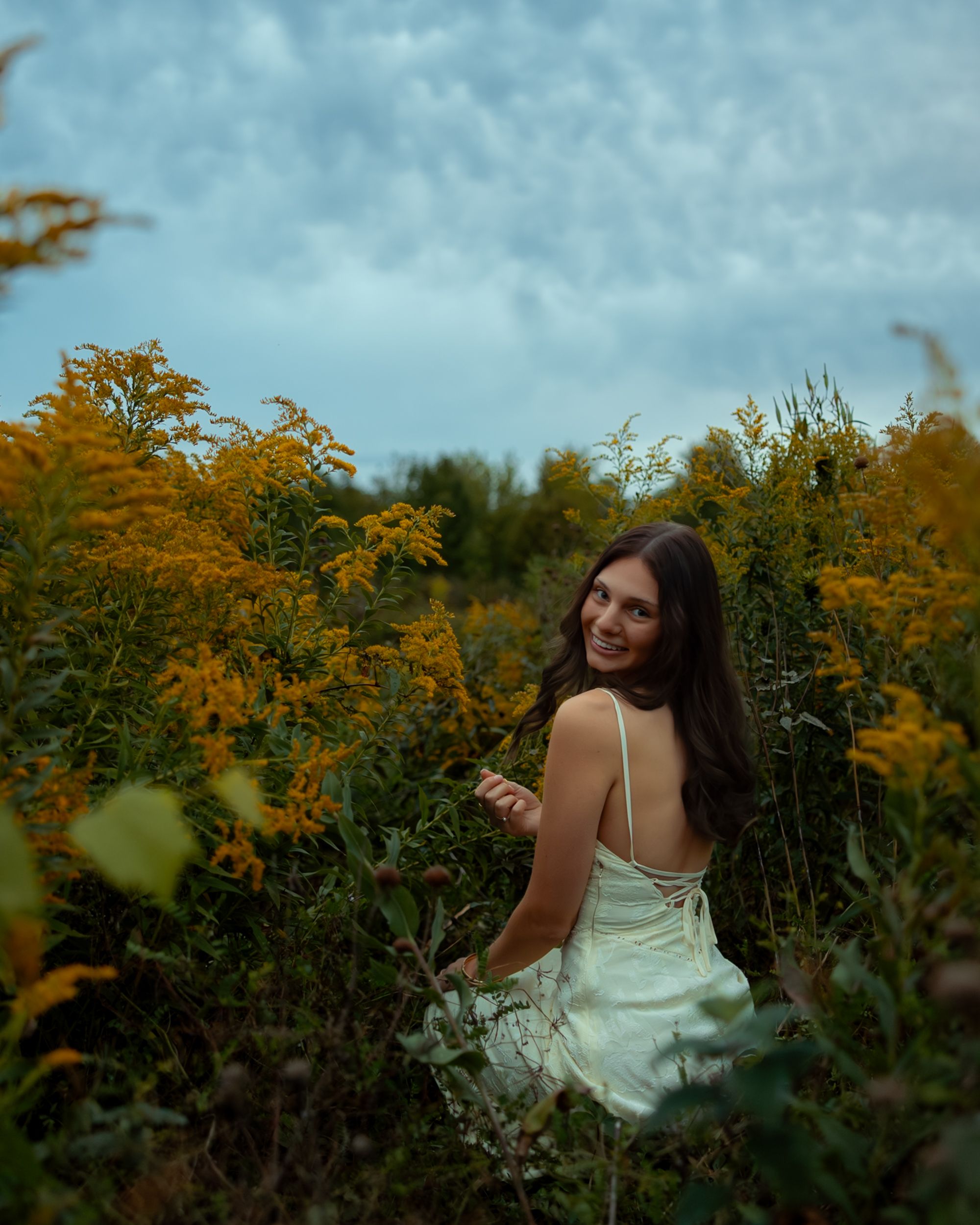 A figure in a flowing white dress poses gracefully among blooming yellow wildflowers against a moody blue sky.
