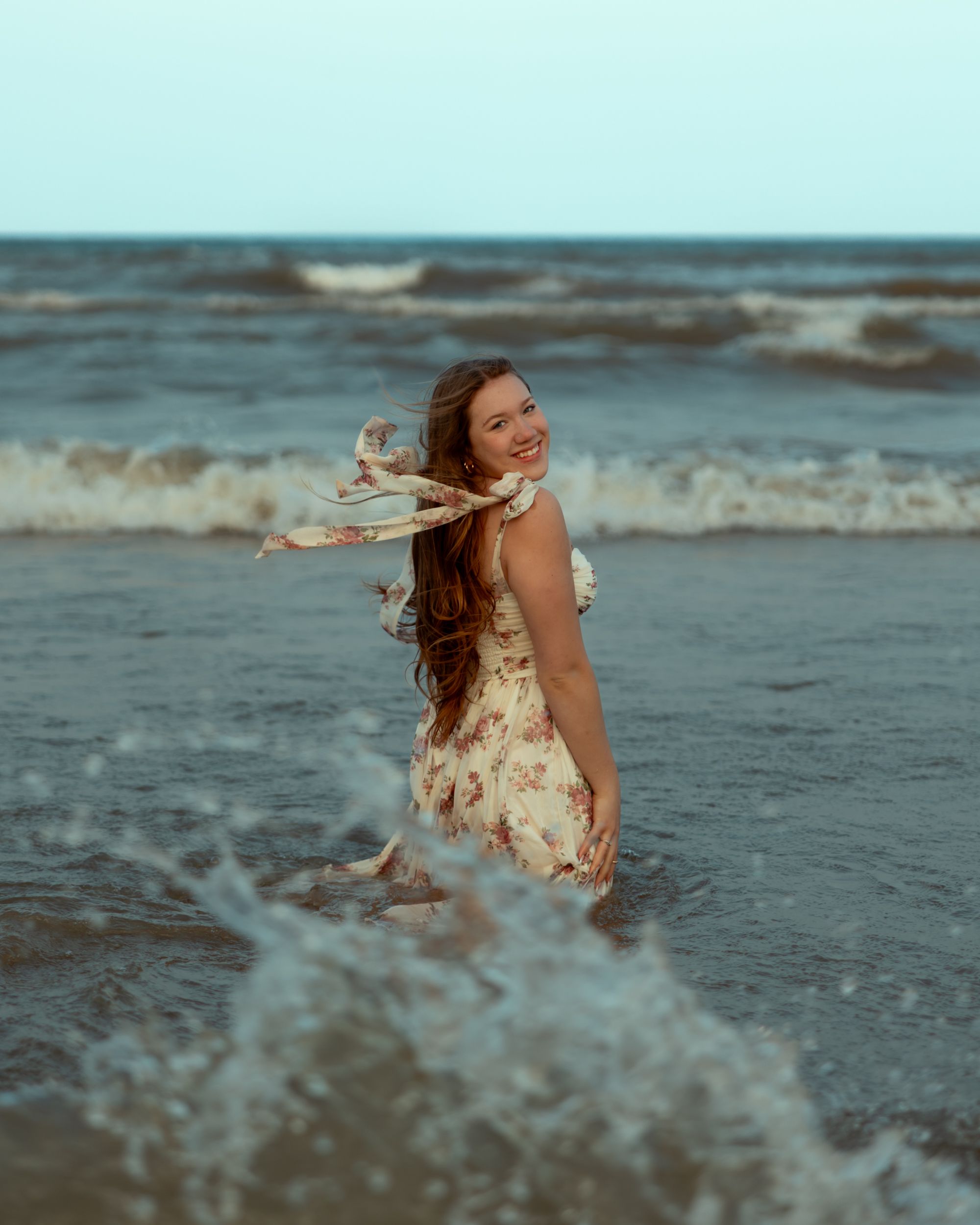 A person in a floral dress dances and twirls gracefully along the beach shoreline at sunset.